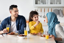 Cute Little Arab Girl Playfully Feeding Mom While Having Breakfast With Parents In Kitchen, Happy Muslim Family Of Three Sitting At Table And Enjoying Tasty Morning Meal Together, Closeup Shot
