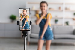Young Blogger Concept. Cheerful teen girl dancing at camera filming video using phone on tripod at home, creating her trendy content on a mobile app to share on social media, selective focus on screen