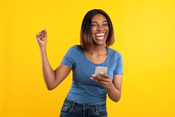 Happy Black Lady Holding Phone Gesturing Yes Winning Mobile Game Over Yellow Background. Studio Shot Of African Woman Celebrating Great News Posing With Cellphone