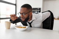 Hurry To Work. Portrait of black business man having breakfast in kitchen at home, African American male in glasses wearing suit while eating food, sitting at dining table, rushing to office