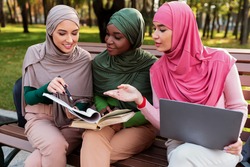 Group Of Modern Muslim Students Ladies Learning Reading Books Sitting On Bench Outside. Islamic Females Learn Together Using Laptop Discussing Something In University Campus Park