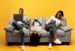Gadgets addiction. Young black family of three holding and using different electronic devices while sitting on sofa on yellow background. Parents and their daughter with modern gadgets