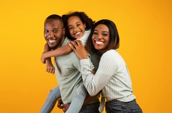 Time with family. Overjoyed african american family laughing and posing to camera isolated over yellow studio wall. Cheerful father carrying his daughter on back