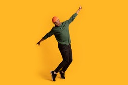Cheerful young african american man in orange hat standing on tiptoes over yellow background, full length shot of happy millennial black hipster guy dancing and having fun in studio, copy space