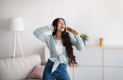 Beautiful Indian lady with headphones dancing and singing, using hairbrush as microphone at home. Millennial woman moving to her favorite song, enjoying music, pretending to be popular star