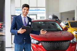 Handsome middle-eastern arab man sales manager showing brand new automobile in luxury auto showroom, holding chart, giving information about nice red automobile, copy space. Car retail concept