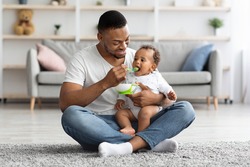 Caring Black Dad Feeding His Adorable Infant Baby From Spoon While Sitting Together On Carpet In Living Room, Young African American Father Enjoying Takic Care About Little Child, Copy Space