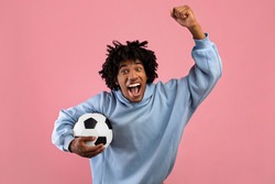 Happy black teenage guy with soccer ball rooting for his favorite football team on pink studio background. Excited African American sports fan celebrating victory, shouting GOAL