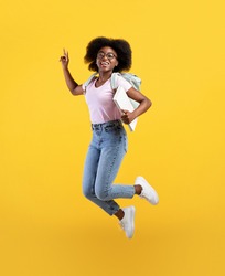Enjoying student life. Full length portrait of african american lady jumping, wearing backpack and holding notepads over yellow studio background, crop