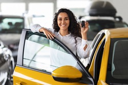 Happy brunette woman buying new cute car, raising hand up with automatic key and smiling, showroom interior, copy space. Pretty curly lady customer purchasing yellow female auto, getting in