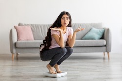 Surprised Indian lady sitting on scales, cannot understand lack of weight loss at home, copy space. Millennial Asian lady shocked over result of slimming diet. Healthy living concept