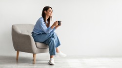 Full length of young woman drinking hot coffee in cozy armchair against white studio wall, banner design with free space. Peaceful lady having relaxing day, chilling on lazy morning