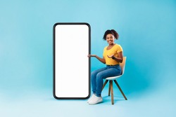 Full length of pretty Afro lady sitting on chair next to big blank smartphone with mockup for mobile app or website design, blue studio background. Space for your online advertisement
