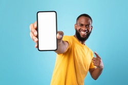 Excited black guy in casual wear pointing at cellphone with empty screen on blue studio background, space for website or mobile app design. Cellphone display mockup. Selective focus