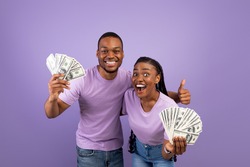 Portrait of overjoyed young African American couple holding and showing bunch of money, celebrating success together, making thumb up gesture, posing over purple studio background, copy space