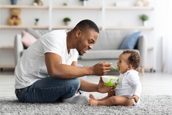 Father's Care. Loving Black Dad Feeding His Cute Baby Son From Spoon At Home, Young African American Daddy Giving Healthy Food To His Little Toddler Child While They Relaxing Together In Living Room