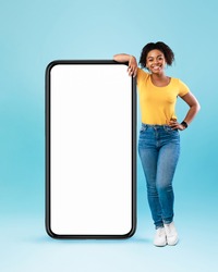Happy black woman leaning on big smartphone with blank white screen, smiling at camera on blue studio background, mockup for mobile app, website, your advertisement design