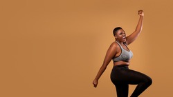 Excited black chubby woman in sportswear dancing, emotional happy lady celebrating success and victory, jumping, cheering and raising clenched fist up, brown studio background, banner, free copy space
