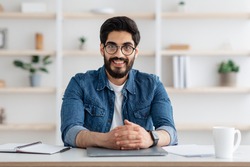 Happy toung arab self-entrepreneur man sitting at desk in home office interior and smiling to camera. Handsome millennial businessman posing at workplace, copy space