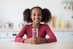 Cheerful african american pretty girl teenager drinking water while sitting at table, smiling at camera, kitchen interior, copy space. Hydration, fresh water drinking for kids, teenagers concept