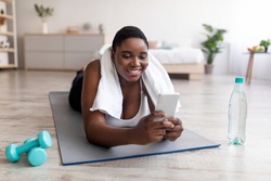 Positive curvy black woman using smartphone during her break from home training, browsing internet, looking up workout video online. Overweight Afro lady checking cellphone after exercising