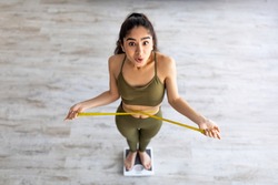 Above view of shocked Indian lady standing on scales, checking her waist measurements, surprised with unexpected results of weight loss program, indoors. Slimming diet and workout concept