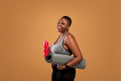 Fitness Concept. Portrait of happy black plus size woman holding water bottle and yoga mat, body positive female smiling and looking at camera standing isolated over brown studio background