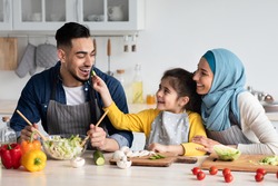 Taste It. Portrait of cute little arab girl feeding her daddy with cucumber slice, happy middle eastern family of three cooking healthy tasty food in kitchen together, making vegetable meal at home