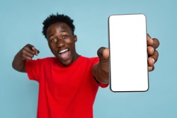 Excited Young Black Man Pointing On Smartphone With Blank White Screen In His Hands, Emotional African American Guy Demonstrating Copy Space For Your Design, Standing On Blue Background, Mockup