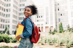Happy African American Student Girl Posing With Backpack Holding Books Smiling Looking Aside Standing Near University Building Outdoor. Modern Education And Studentship Lifestyle