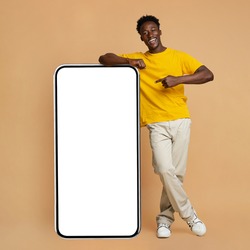 Happy Young Black Man Leaning And Pointing At Big Smartphone With White Blank Screen, Cheerful African Guy Showing Free Copy Space For Your Design, Standing Over Beige Background, Mockup Image