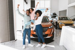 Full length of excited African American family lifting hands up, celebrating new car purchase at auto dealership. Black parents with daughter making YES gesture after buying vehicle at showroom