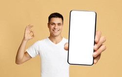 I Recommend. Portrait of smiling man holding smartphone with white blank device screen in hand, showing okay sign gesture, beige studio. Gadget with empty free space for mock up, isolated display