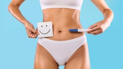 Positive Result. Young woman holding pregnancy test with two stripes and sticker post-it note card with smiley face. Unrecognizable female waiting for a long-awaited planned baby. Closeup cropped view