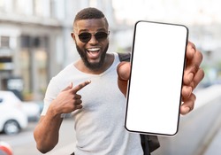Great Offer. Portrait of excited black man standing outdoors holding big phone with white blank screen in hand, showing close to camera pointing at device. Gadget with empty free space for mock up