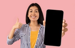 I Recommend. Portrait of smiling asian woman holding smartphone with black blank device screen in hand, showing thumb up gesture, pink studio. Gadget with empty free space for mock up, banner isolated