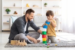Playtime. Cheerful loving father playing colorful blocks with his little son, boy building big tower, spending free time together at home interior