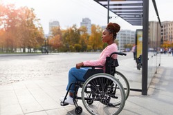 Lack of public transport for disabled people. Young black paraplegic woman in wheelchair feeling upset, waiting on bus stop in autumn, cannot board vehicle suitable for handicapped persons, copy space