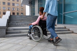 Young black guy pushing wheelchair with handicapped woman, having problem entering building, standing in front of stairs without ramp. Problems with accessible environment for disabled people