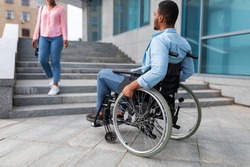 Handicapped people problems. African American disabled guy suffering from lack of wheelchair friendly facilities, looking at woman going down stairs, cannot enter building without ramp