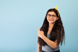 Promotion concept. Cheerful Indian teen girl in casual outfit pointing aside at blank space, recommending something, offering huge sale or discount on blue studio background