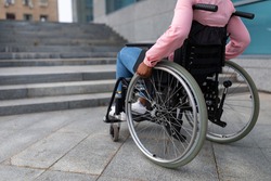 Unrecognizable young black paraplegic woman in wheelchair cannot go up stairs without ramp, feeling limited and unhappy, cropped. Copy space. Disability, environment, mobility and social participation