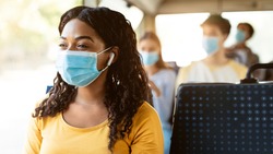 Modern Travels. Portrait of happy smiling black female passenger in medical face mask traveling on public transit, listening to music in wireless earphones looking away at window, thinking, panorama