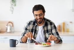 Positive arab guy hipster having healthy breakfast at home, eating fresh vegetables, sausages, drinking coffee, sitting at kitchen table, copy space. Handsome indian man starting day with nice meal