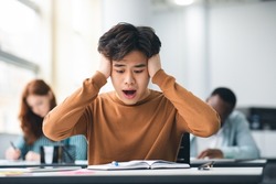 Portrait of shocked asian freshman sitting at table in classroom, grabbing clasping his head in horror and panic, looking at hard exam questions or test result, unhappy about deadline or low score