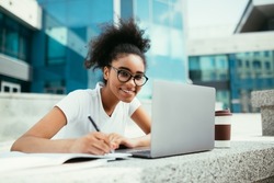 E-Learning. Cheerful African Student Girl Taking Notes At Laptop Learning Distantly Sitting Outdoor Near Urban College Building, Wearing Eyeglasses. Modern Online Education