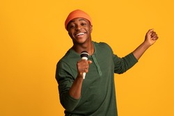 Cheerful Black Millennial Guy Holding Microphone And Singing At Camera, Young Joyful African American Hipster Man Enjoying Karaoke, Having Fun While Standing Over Yellow Studio Background, Copy Space