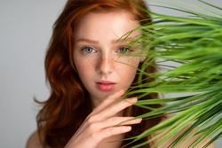 Headshot Of Young Lady With Red Hair Posing Over Gray Background With Green Plant On Front. Selective Focus On Beautiful Female Face. Beauty And Natural Cosmetics Concept