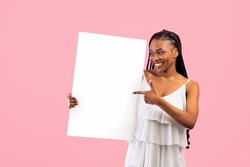 Joyful black woman with Afro bunches pointing at empty banner with space for design on pink background. Beautiful African American lady showing blank poster, mockup for advertisement