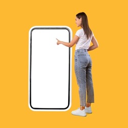 Full Body Length Back View Of Woman Using Big Smartphone With Blank White Screen And Touching Display Panel With Finger, Cheerful Lady Standing On Yellow Background, Ordering Food Delivery, Mock Up